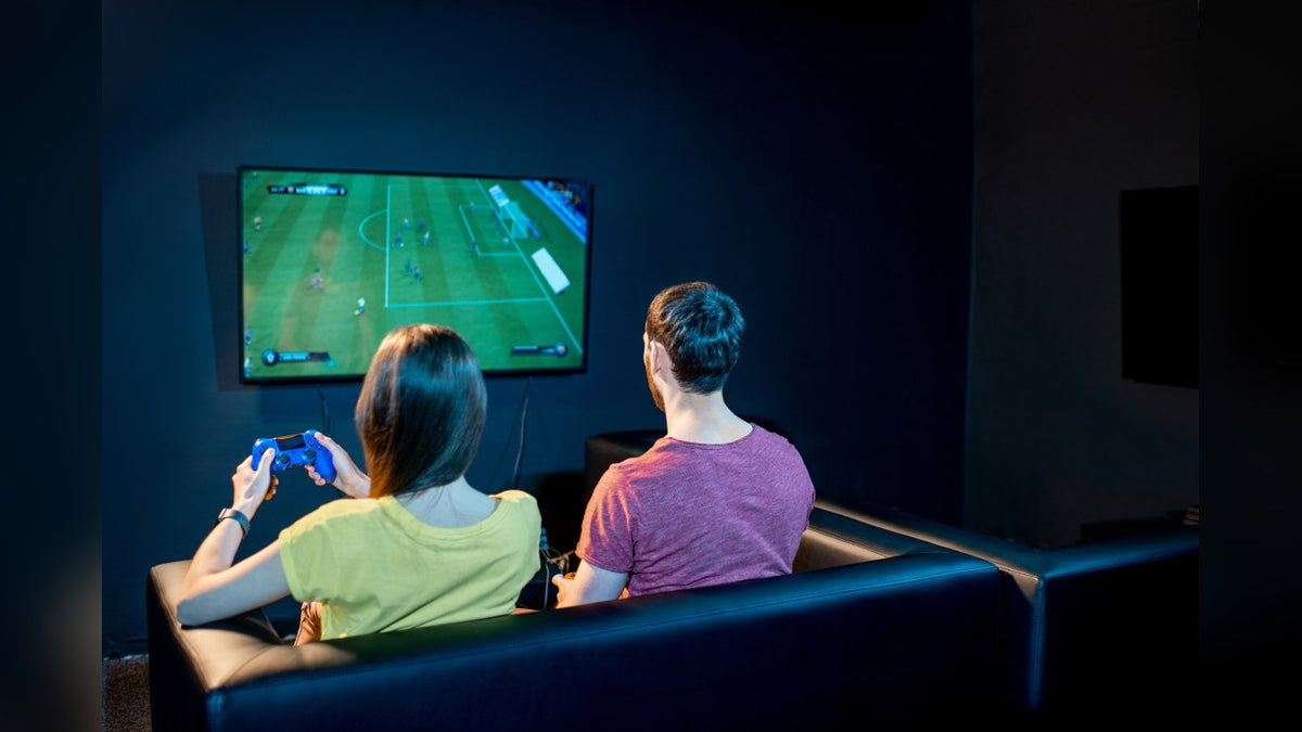 Two people on a leather couch playing a soccer video game on a wall-mounted TV with a console controller