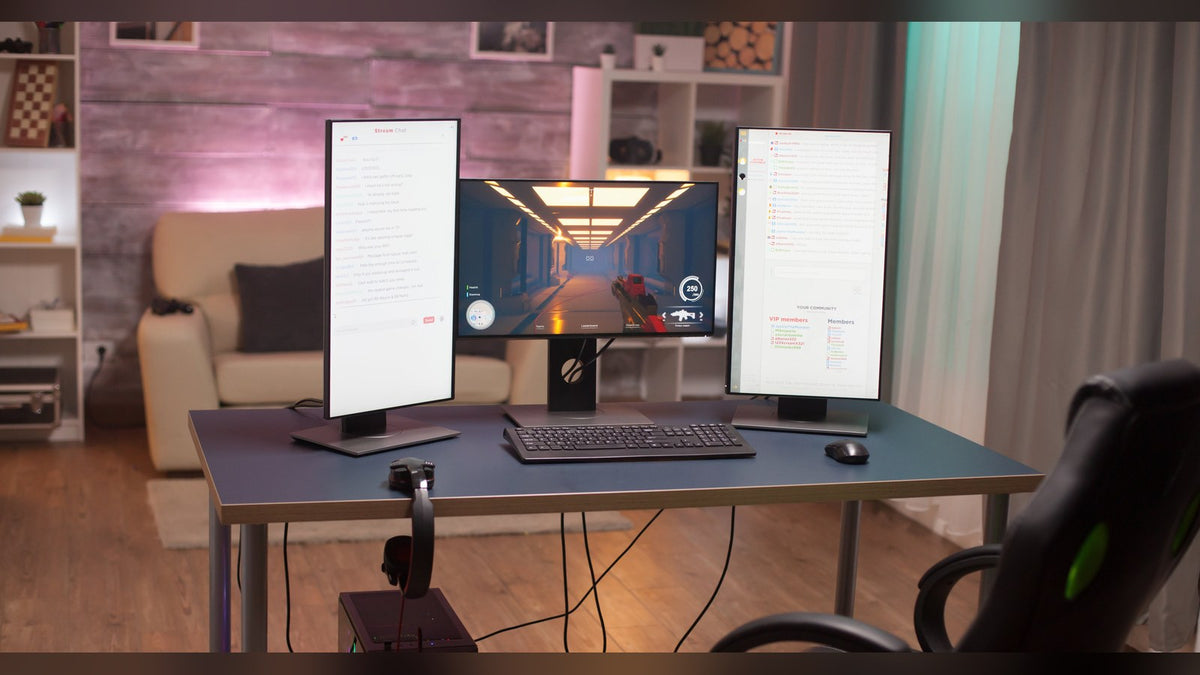 Home desk with a horizontal monitor running a shooter game, flanked by two vertical monitors showing chat windows