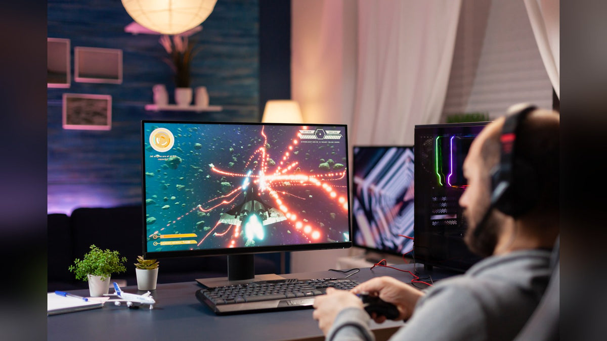 Gamer in a headset playing a space shooter on a monitor, with a second screen and RGB-lit PC tower on the desk