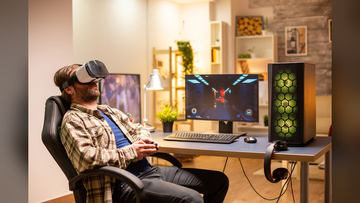 Man in a VR headset holding a controller at a desk beside a monitor and a mesh PC tower with green-lit fans
