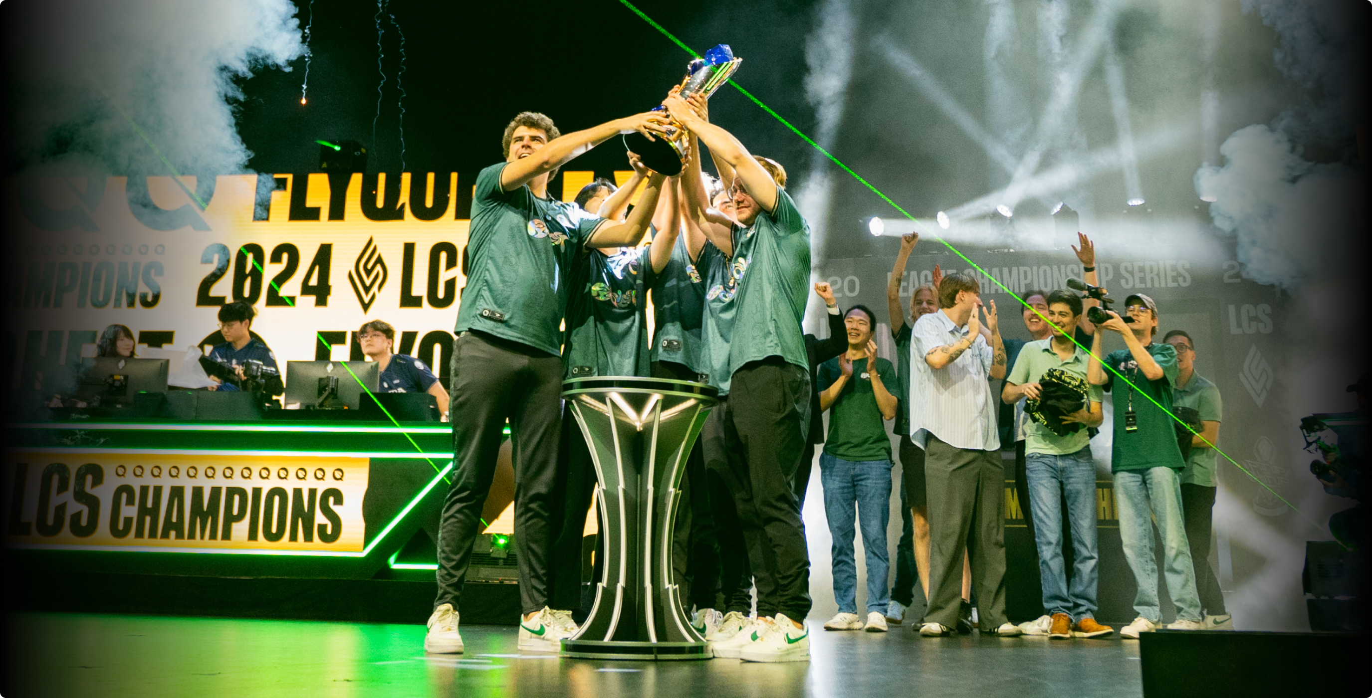 A team of esports players celebrates on stage, holding up a trophy together. Behind them, large screens display 2024 LCS Champions. Staff and fans cheer in the background amid bright lights and green laser effects.