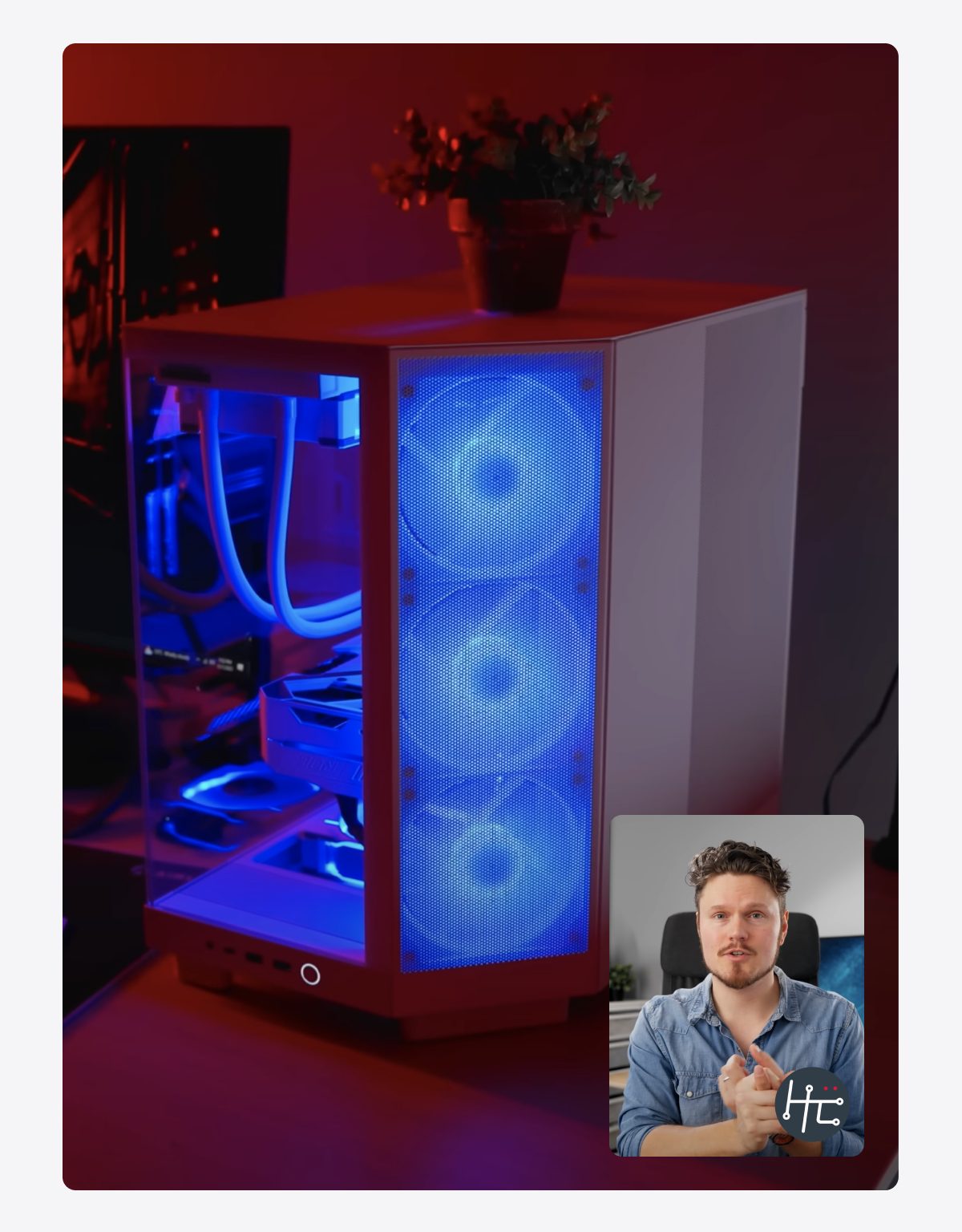 A white gaming PC with blue LED fans and visible liquid cooling sits on a desk. A small video overlay in the corner shows a man with a beard speaking in front of a plant and shelves.