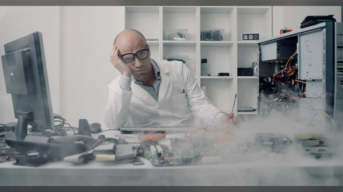 Frustrated man in a lab coat at a desk of scattered PC parts, next to an open PC tower with smoke rising