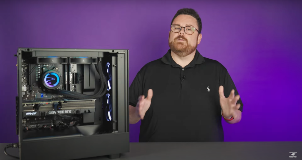 A man with glasses and a beard stands next to an open desktop computer with visible components, gesturing with his hands, against a solid purple background.